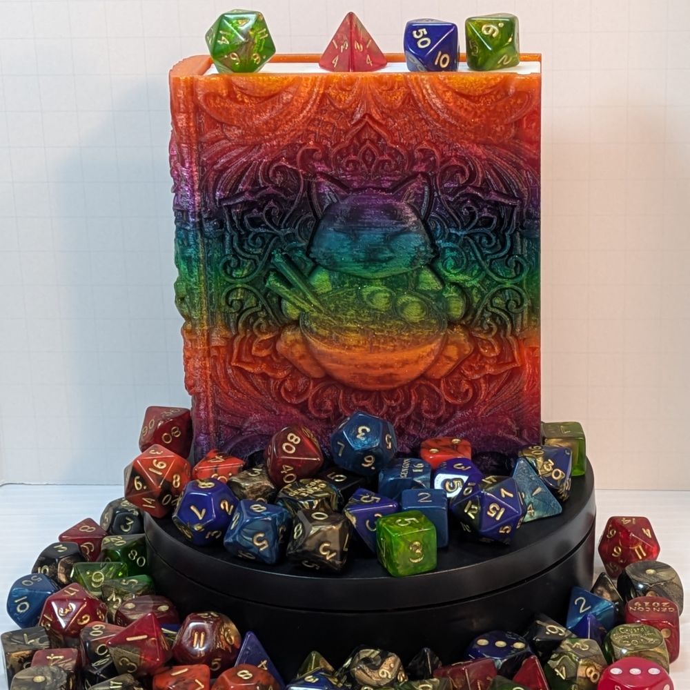 A dice box shaped like a tome. It's surround by colorful TTRPG dice. The box is printed out of a shiny rainbow gradient filament, and it has a happy cat sitting behind a bowl of ramen.