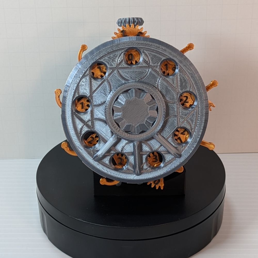The side of the dice box. It's shaped like a large pocket watch, with knotwork on the side and gears and levers sticking out of the lid to change numbers on the counter. The box itself is a metallic silver, and the switches are bronze with black numbers.