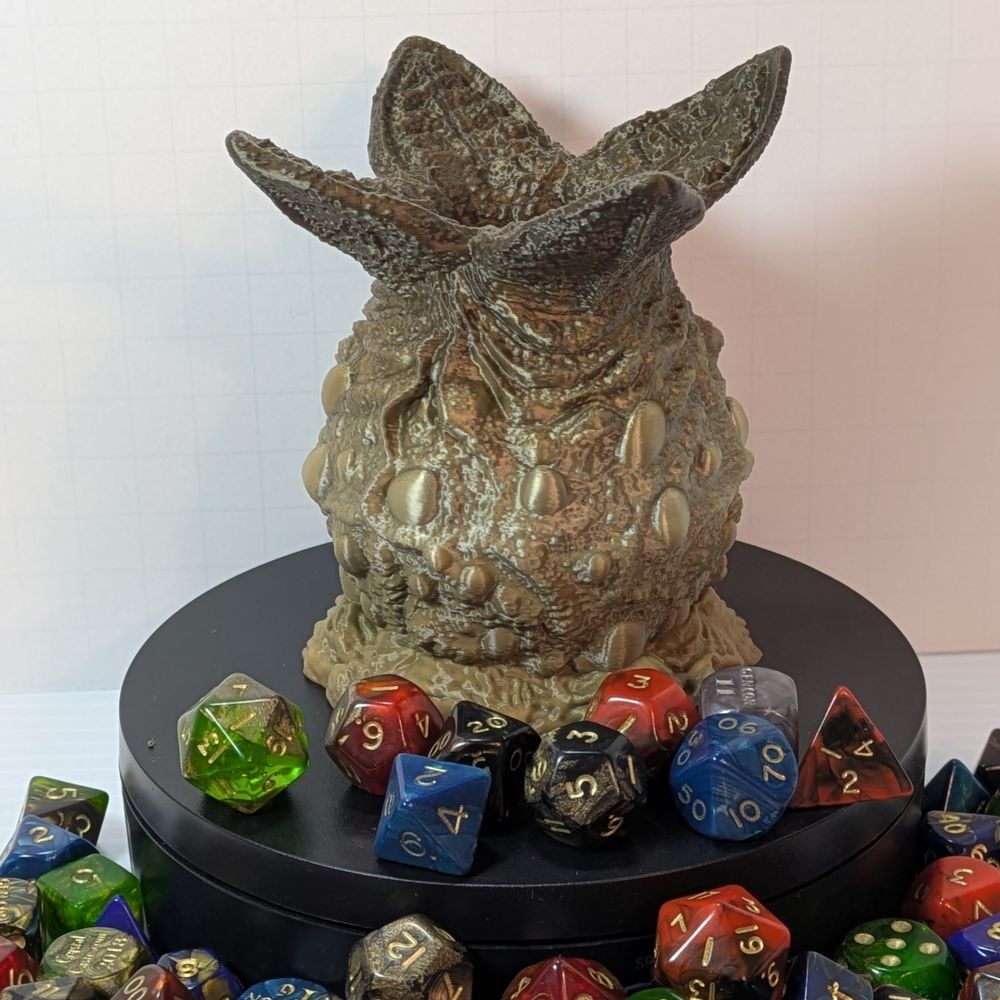 A 3D printed dice holder shaped like an egg from an alien. This one is in a green gradient filament, and there are four wedges at the top spread out, like the egg has been opened. The egg is lumpy and kind of gross looking.