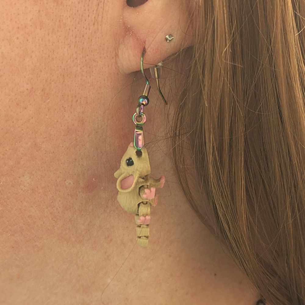 An earring in the shape of a tiny cartoonish mouse. It is 3D Printed, in color, a light brown with black and white eyes and pink in the ears, nose, and toes. The "fish hook" style earring is clamped in the mouse's mouth, and they're displayed on an ear.