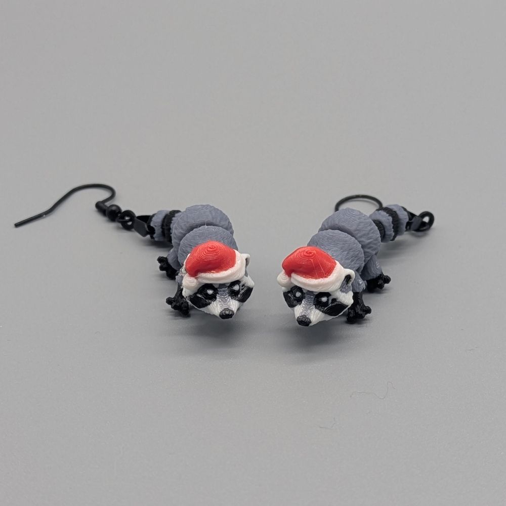 A pair of 3D printed earrings of raccoons wearing Santa hats.
