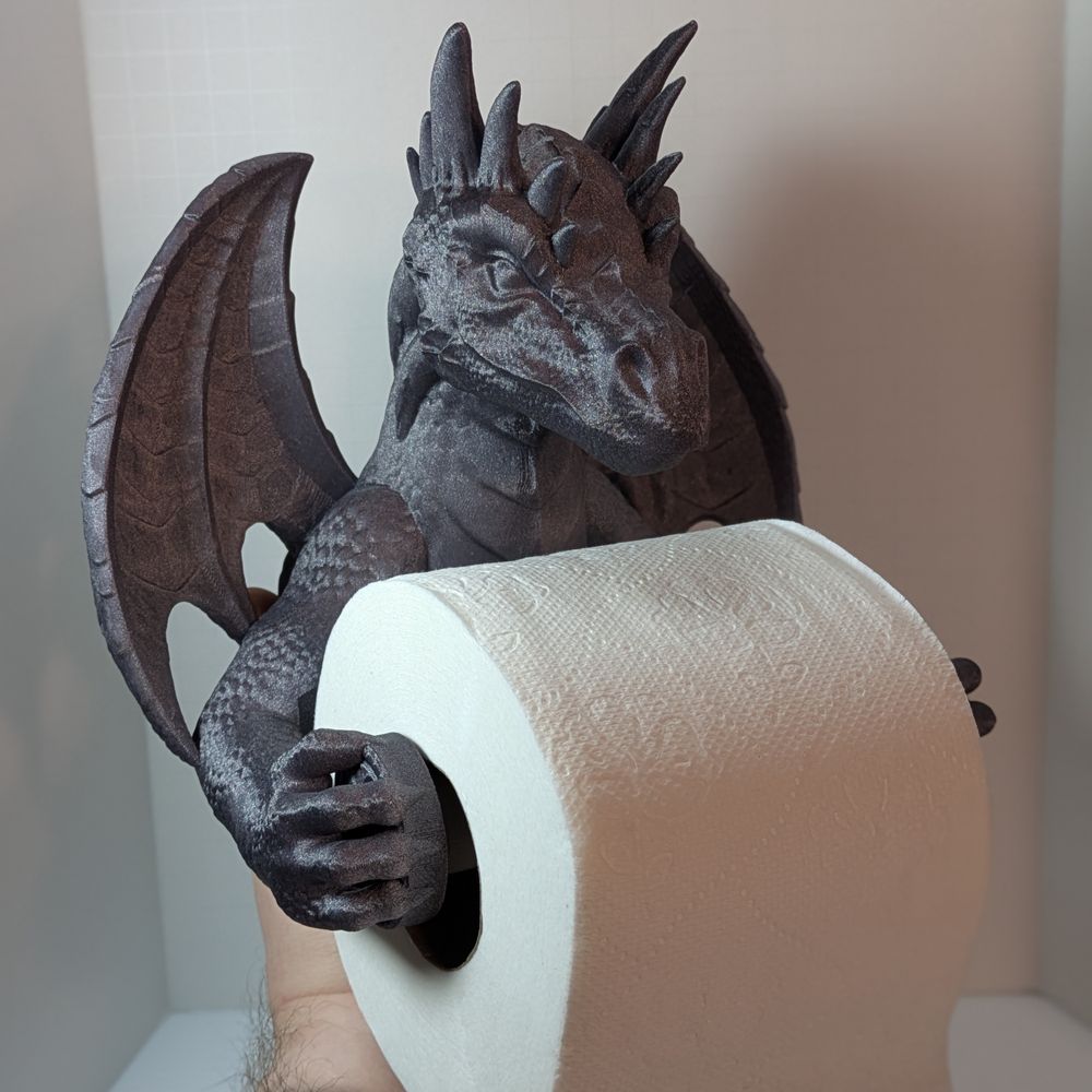A 3D printed toilet paper holder in the shape of a dragon. It's a dark silver color, and it's being held up with toilet paper on the roll. The roll is held between the dragon's outstretch arms, with the spiked head looking over it.