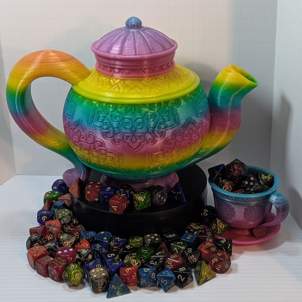 A 3D printed teapot, tea cup, and saucer. The teapot is on a black circular stand, surrounded by colorful TTRPG dice. The cup and saucer are sitting underneath the spout, and the cup is filled with dice, as well.