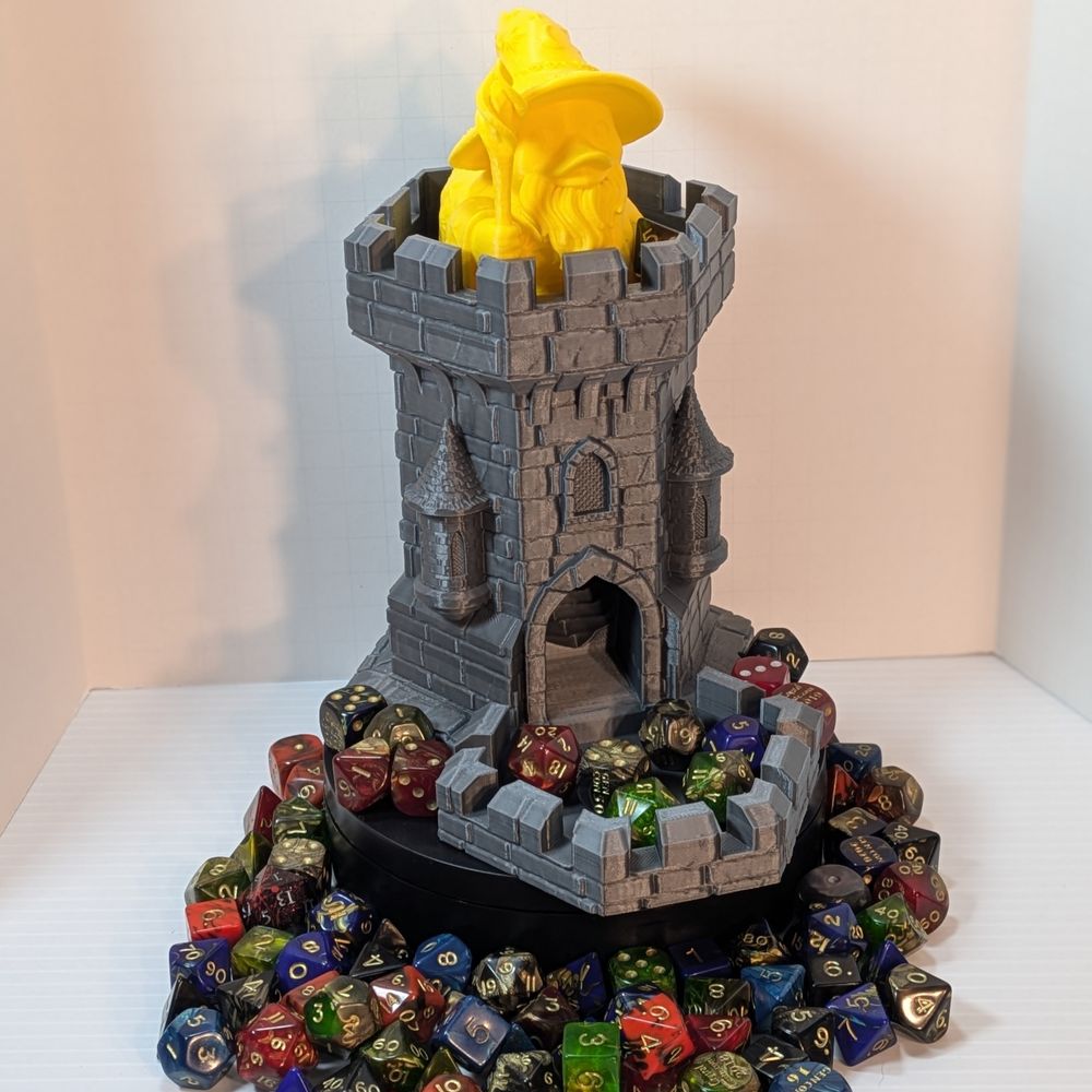 A 3D printed dice tower in the shape of a castle tower. It's a metallic steel color, surrounded by colorful TTRPG dice. At the top is a yellow ducks, dressed like a wizard.