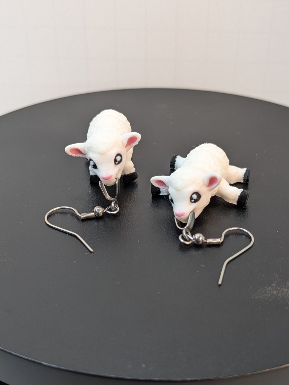 A pair of 3D printed earrings in the shape of lambs. One is standing on four legs looking at the camera, the other is laying on it's stomach with the legs splayed out at the sides. They both have silver earring hooks clasped in their mouths.