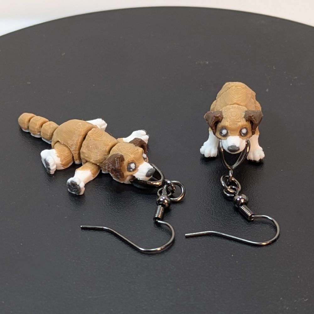 A pair of 3D printed earrings, shaped like dogs. It looks like it could be a lab mix, holding the dark metal fish hook earrings in it's mouth. It has dark brown ears, a tan and white coat, and a black nose and paw pads. One is standing, one is laying down