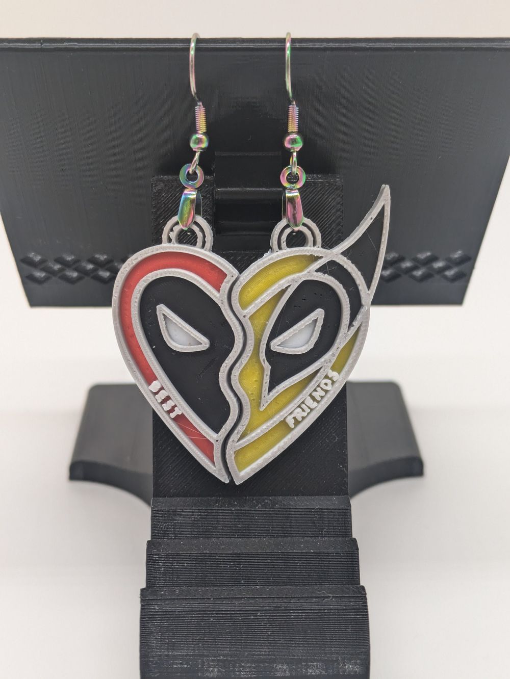 A pair of earrings that look like they're the "Best Friend" necklace of a heart cut in half. One half is a Deadpool mask, the other Wolverine.