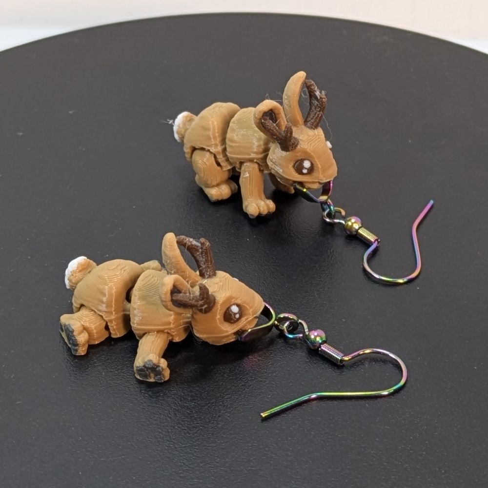 Side view of a pair of 3D printed earrings, shaped like Jackalopes. They are light brown, with dark brown prongs, and they're attached to rainbow colored stainless steel earring hooks.