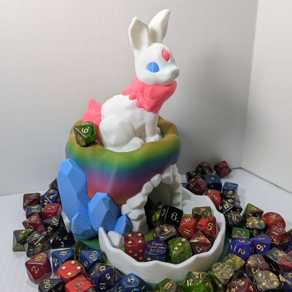 A 3D printed dice tower, with a white tray and blue crystal rocks surrounding a rainbow base, which has a white bunny with pink markings and blue eyes sitting on top. It's surrounded by colorful TTRPG dice.