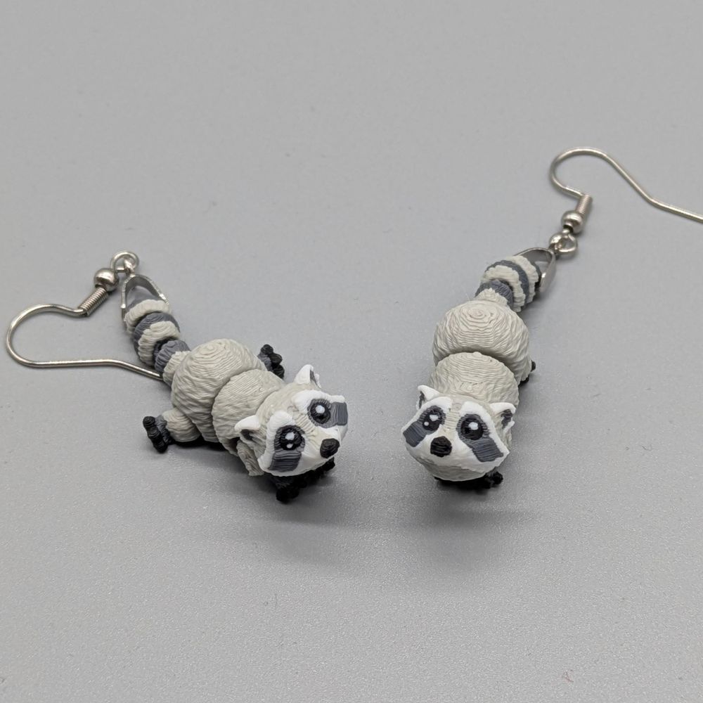 A pair of raccoon earrings, 3D printed in color. They have some light articulation, and one is standing while the other is laying down.