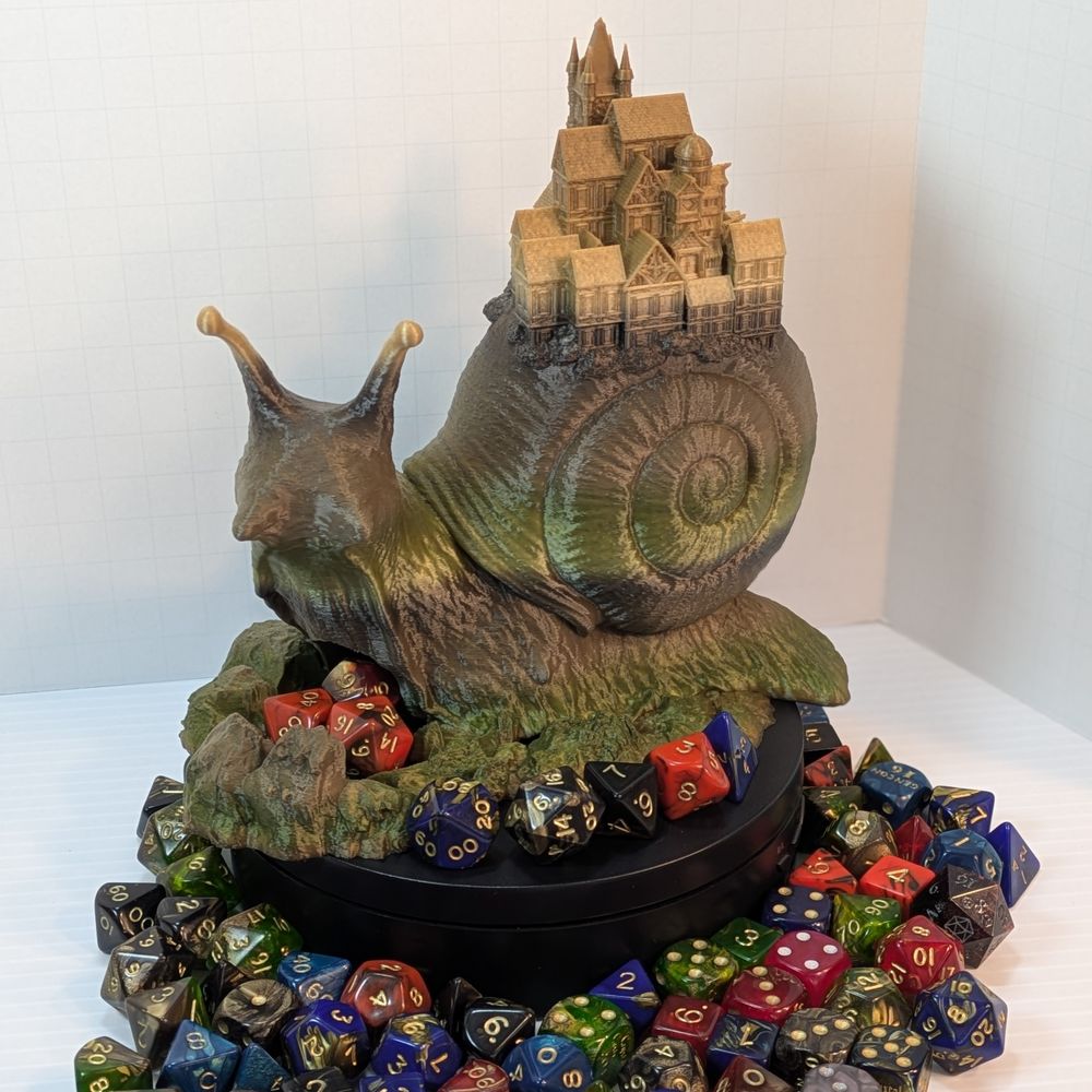 A 3D printed dice tower in the shape of a snail, with a small town on the back of its shell. The snail is printed in a green and brown gradient, and it's surround by colorful TTRPG dice.