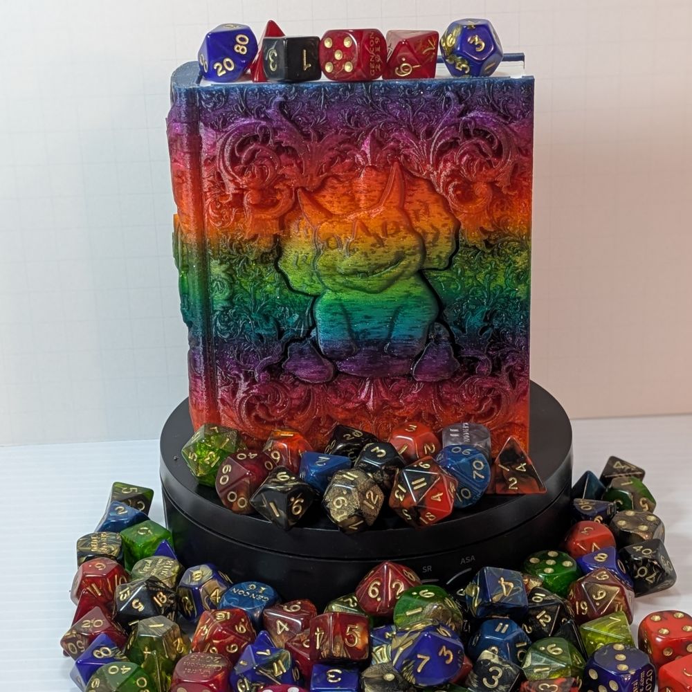 A colorful 3D printed "tome" surrounded by TTRPG dice. The tome has a pudgy baby triceratops sitting on the front of it.