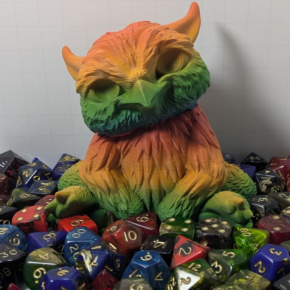 A 3D printed baby owlbear, printed in a rainbow gradient. It is sitting, with their front legs/arms draped in front of them, their head cocked to the side curiously. They are surrounded by colorful TTRPG dice.