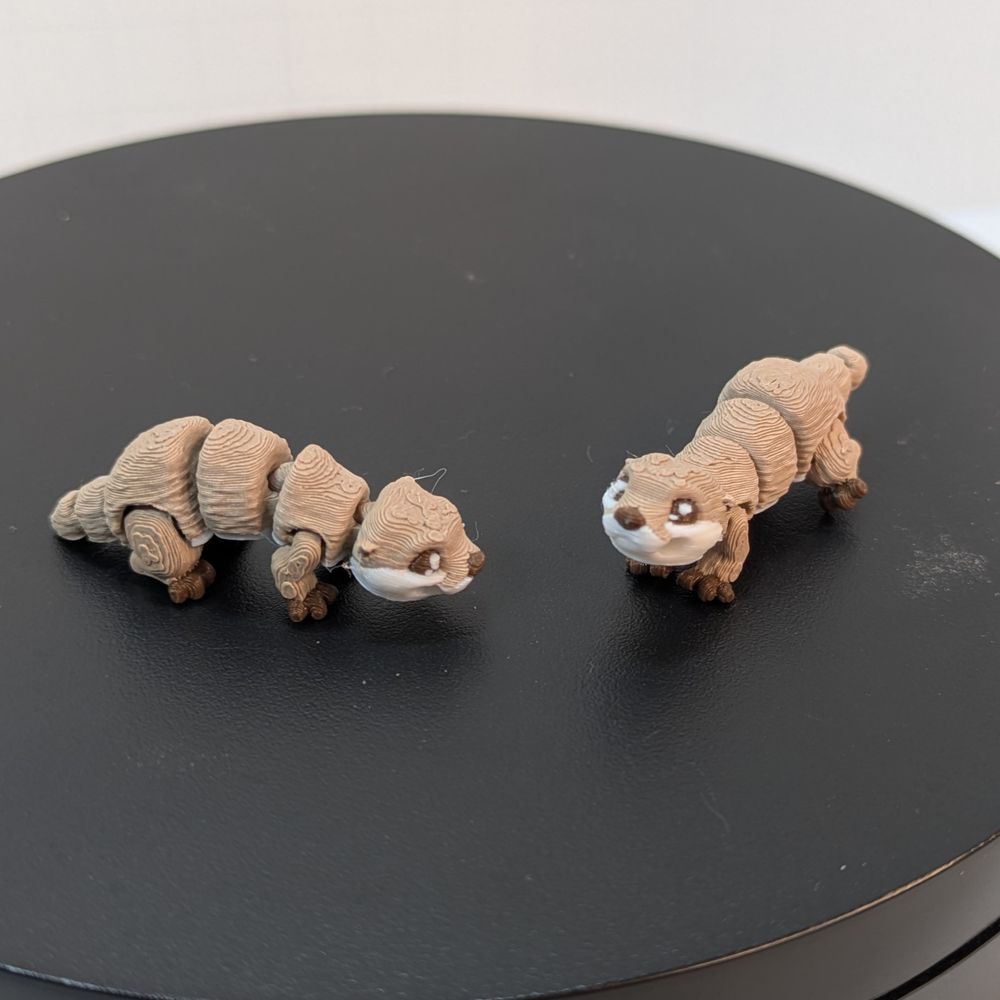 A pair of 3D printed River Otters. They have a hole in the sides of their mouths to hold the earring hook, and they're tan and white in color.