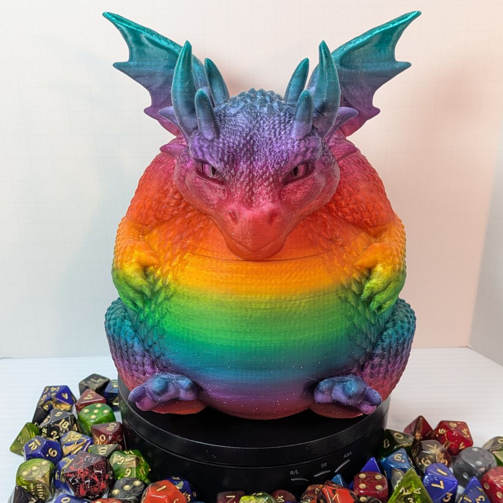 A 3D printed container of a very rotund dragon. It's sitting with is back feet sticking out from under its stomach, and its front paws grasping the sides of its belly. It has tiny wings and a mischievous grin on its face.  