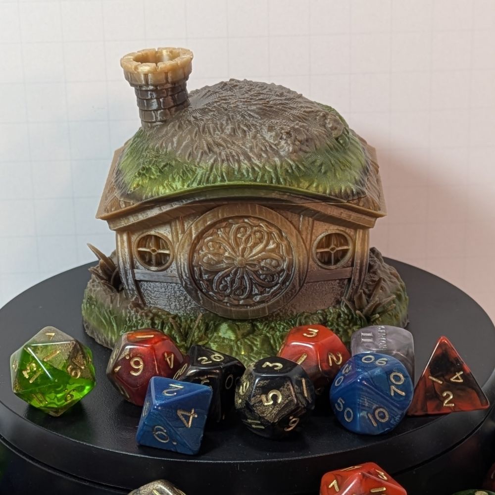 A 3D printed dice vault in the shape of the home of a halfling. It's got grass on the roof, a round chimney, and a round door. The top comes off to put the dice in.