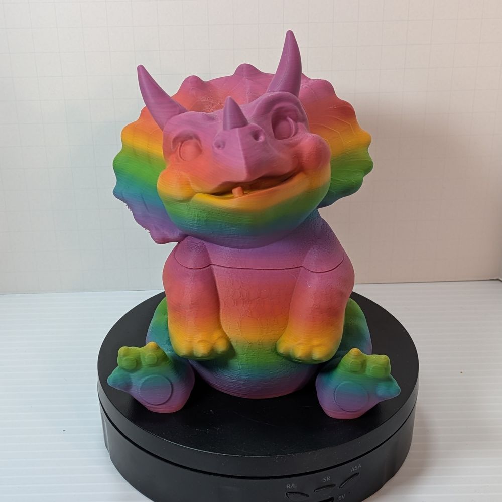 A chunky little baby triceratops, 3D printed out of a bright rainbow PLA plastic. There is a seam at the shoulders where you can lift the head off and use it as a dice vault.