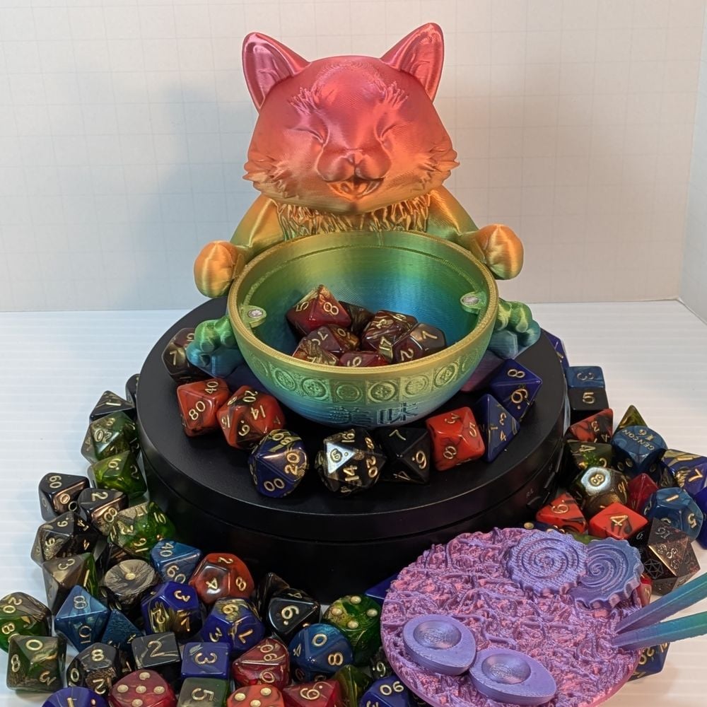 A 3d printed dice vault shaped like a chubby cat sitting behind a bow of ramen. The ramen and chopsticks are removed, showing a set of dice inside the bowl. It is surrounded by a colorful array of dice, and it rests on a black platform.