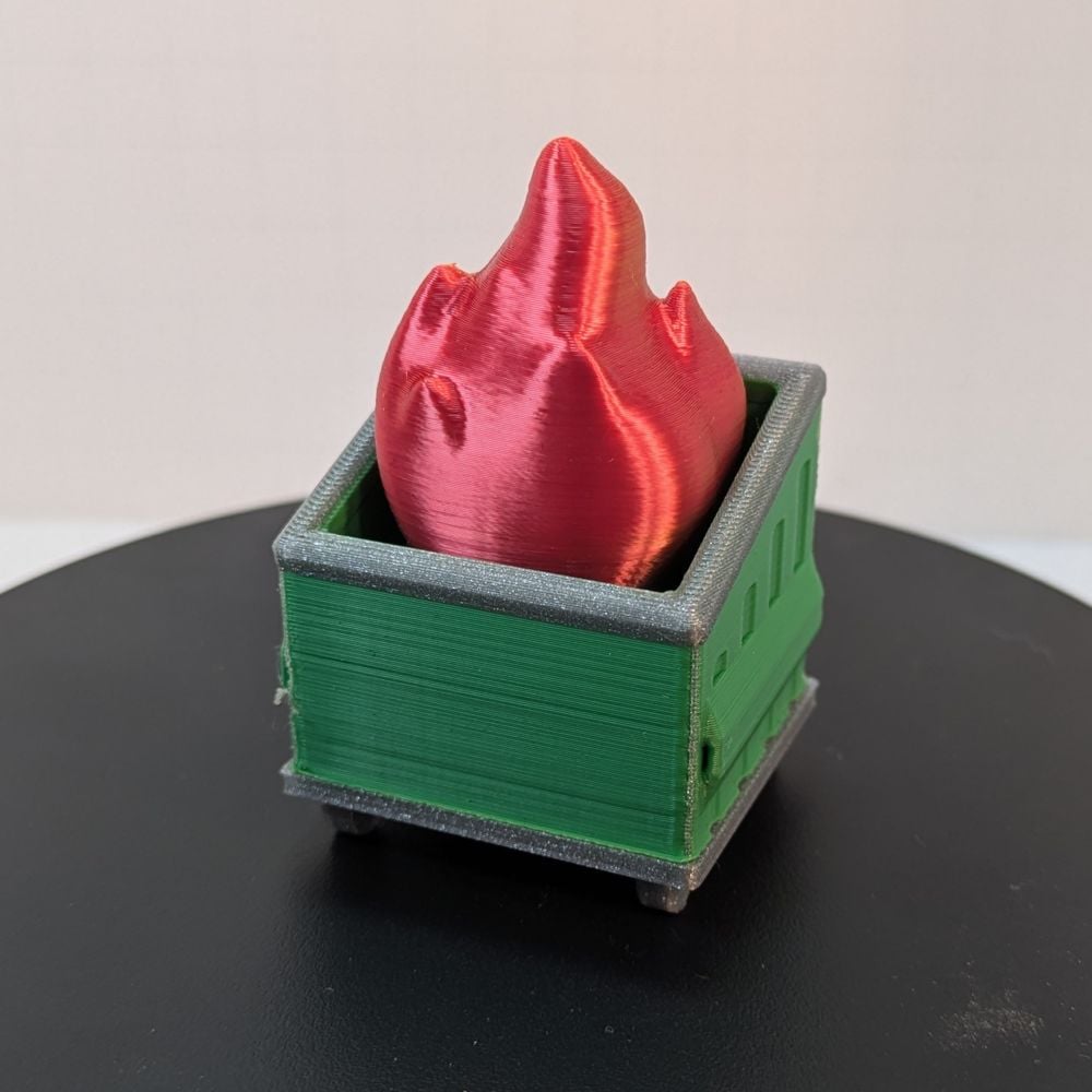 A 3D printed dumpster fire. The dumpster is green with a silver base and rim around the top, and the fire coming out the top is a shiny red with yellow accents.