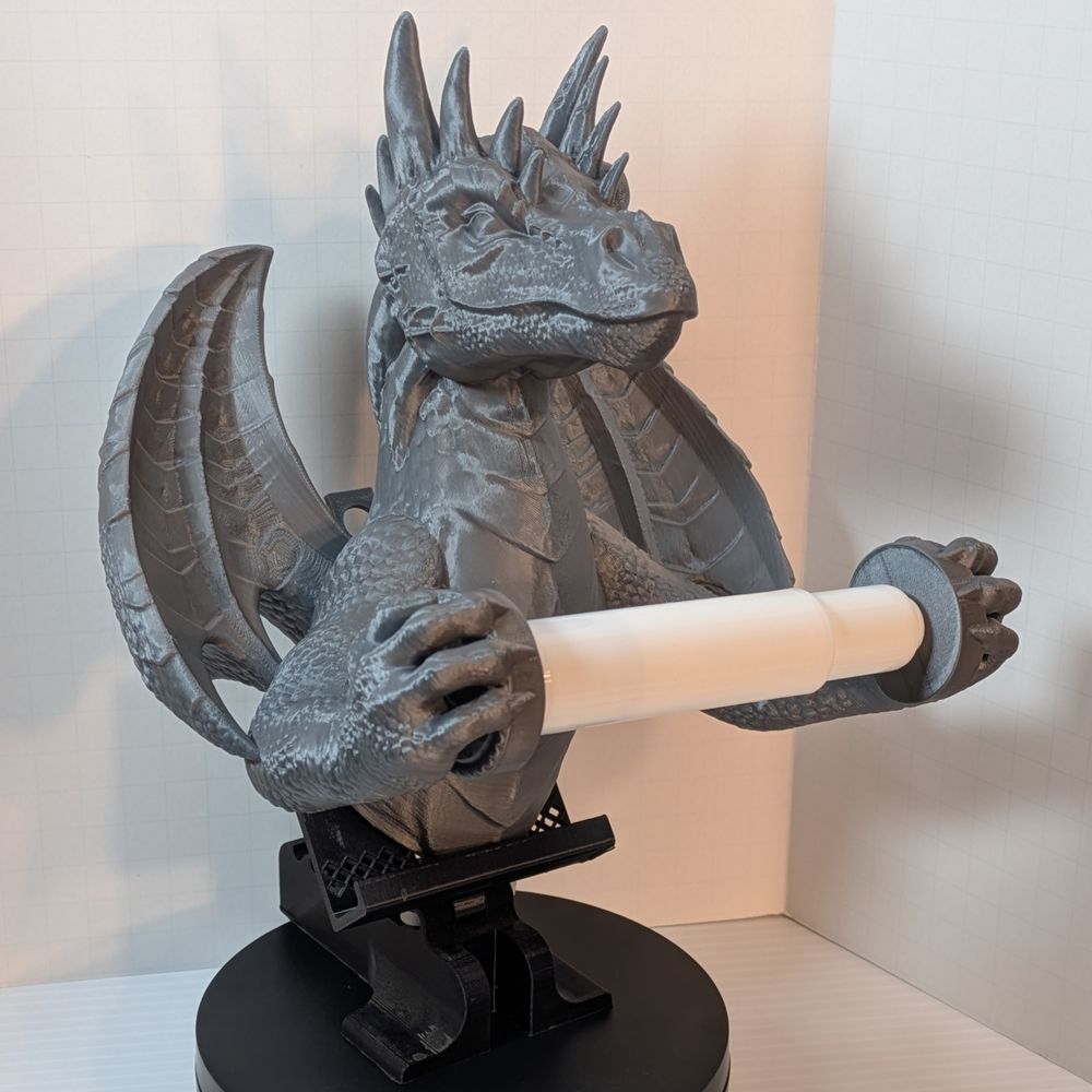 A 3D printed toilet paper holder in the shape of a dragon. It's a dull Steel color, and its front claws are holding the spindle for the toilet paper roll out away from its chest.